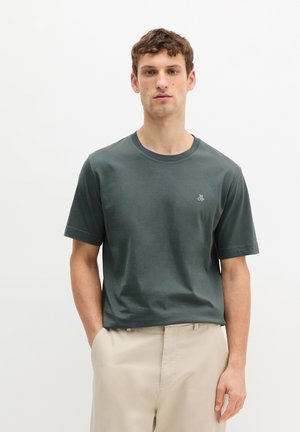 Homme debout les mains dans les poches, portant un T-shirt à manches courtes vert foncé et un pantalon beige, sur fond blanc uni.