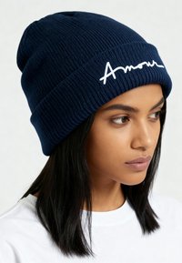 Bonnet en maille côtelée marine avec le texte "Amour" écrit à la main en blanc. Texture douce avec un ajustement près du corps, idéal pour la chaleur par temps froid.