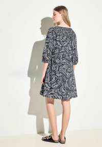 Vestido negro con un patrón de paisley blanco, mangas tres cuartos y un corte holgado, con un escote y dobladillo fruncidos por encima de la rodilla.