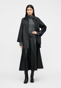 Γυναίκα με μαύρο υπερμεγέθη μπουφάν, γκρι πουλόβερ με λαιμόκοψη, μαύρη μακριά φούστα και μαύρα ψηλοτάκουνα μπότες, στέκεται μπροστά σε απλό φόντο.