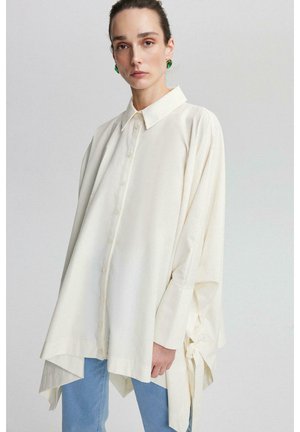 Camicia bianca oversize con colletto a punta, bottoni frontali e maniche lunghe. Presenta dettagli con fiocchi ai lati e una vestibilità rilassata.