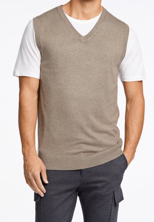 Man wearing beige V-neck sleeveless sweater over white T-shirt with dark cargo pants, standing with one hand in pocket.