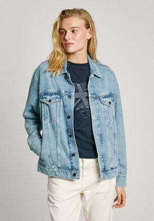 Chaqueta de mezclilla azul clara con dos bolsillos frontales, cierre de botones y detalles desgastados, llevada sobre una camiseta oscura y pantalones blancos.