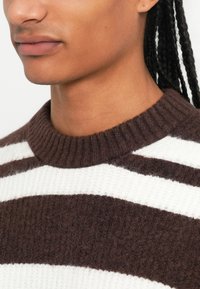 Un pull rayé marron et blanc, avec un col rond côtelé, une texture douce, en matière tricotée, présentant des rayures larges et étroites alternées.