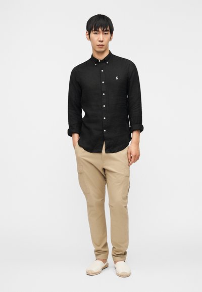 Bărbat purtând o cămașă neagră cu mâneci lungi și nasturi, pantaloni cargo bej și pantofi albi slip-on, stând cu mâinile în buzunare pe fundal alb.