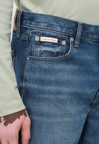 Primer plano de un bolsillo de jeans de denim azul con la etiqueta de Calvin Klein y soldaduras visibles, al lado de una mano con vitiligo y una camiseta de manga larga verde claro.