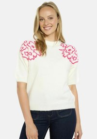 Maglione bianco a maniche corte con ricami floreali rosa sulle spalle, tessuto morbido, vestibilità rilassata e scollatura rotonda, abbinato a jeans scuri.