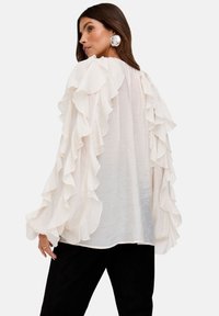 Fließende weiße Bluse mit Rüschenärmeln und strukturiertem Stoff. Der Rücken ist leicht durchsichtig, kombiniert mit schwarzen, maßgeschneiderten Hosen.