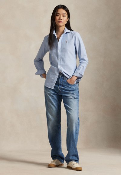 Ung kvinde står med hænderne i lommerne iført en lyseblå skjorte med knapper, blå jeans og hvide sneakers mod en ensfarvet baggrund.