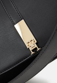 Gros plan d'un sac à main en cuir noir avec un fermoir rectangulaire doré arborant des initiales entremêlées TH sur le rabat.
