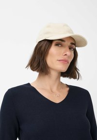 Beige Baseballkappe mit strukturiertem Schirm, gebogenem Visier und glatter Textur, getragen mit einem navyblauen V-Ausschnitt-Pullover und einer schlichten Halskette.