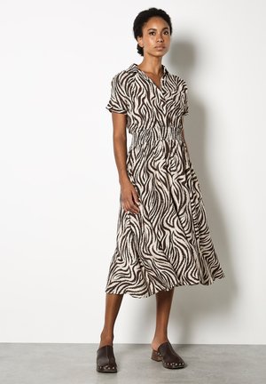 Robe à manches courtes en tissu beige avec un motif zèbre noir en vagues, présentant une taille froncée et une jupe fluide. Associée à des sabots marron.