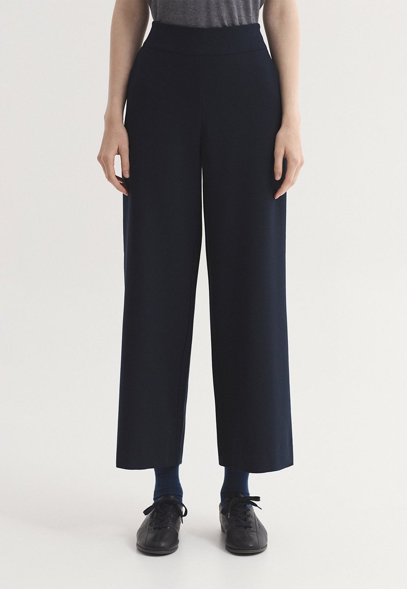 Personne portant un pantalon noir à jambes larges, des chaussettes foncées, des chaussures noires à lacets et une chemise grise rentrée dans le pantalon, se tenant devant un fond blanc uni.