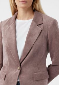 Corduroy blazer in subtiele mauve, met gelapte revers, een enkele knoopsluiting en twee voorzakken. Gemonteerd over een wit T-shirt.