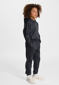 Mörkgrå huvtröja och byxor i slät tyg med elastiska muddar och midjeband; kombinerad med vita sneakers med subtila detaljer.