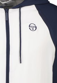Felpa bianca con zip e dettagli blu navy, dotata di cappuccio regolabile tramite coulisse e logo sul petto. Il tessuto presenta una consistenza liscia.
