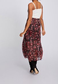 Jupe maxi en tissu noir avec un imprimé floral rouge, dotée d'un design à volants et d'une silhouette fluide. Portée avec un débardeur blanc et des bottes noires.