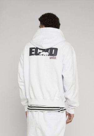 Osoba s kučeravými vlasmi nosiaca bielu mikinu s kapucňou s čierno-hnedým logom "Ecko unltd." a grafikou nosorožca na chrbte.