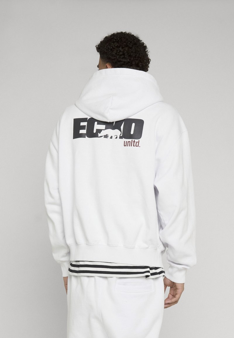 Personne aux cheveux bouclés portant un sweat à capuche blanc avec un logo noir et marron "Ecko unltd." et un graphisme rhinocéros dans le dos.