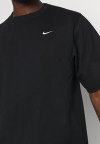 Camiseta negra de manga corta con el logo de Nike en blanco en el pecho izquierdo, llevada por una persona de tono de piel oscura.