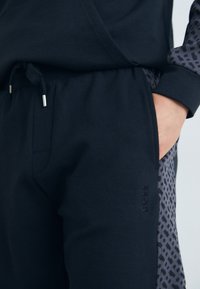 Black joggers made of soft fabric feature a drawstring waistband, side pockets, and a patterned gray side panel with embossed logo detail.