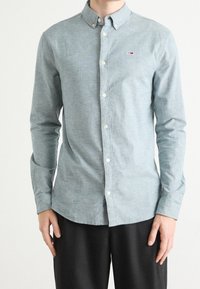 Mies, joka on pukeutunut vaaleansiniseen napitettavaan pitkähihaiseen paitaan, jossa on pieni logo rinnassa, ja mustiin housuihin, seisoo tasaisella taustalla.