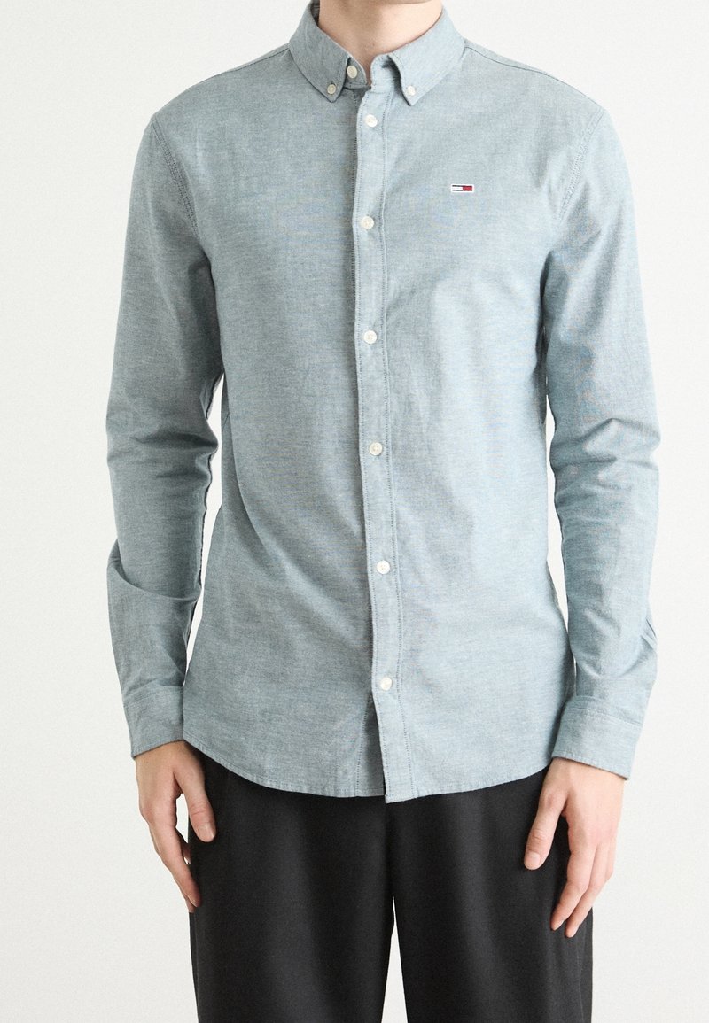 Mies, joka on pukeutunut vaaleansiniseen napitettavaan pitkähihaiseen paitaan, jossa on pieni logo rinnassa, ja mustiin housuihin, seisoo tasaisella taustalla.