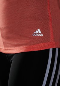 Rode textuur atletische shirt met klein wit Adidas logo, gecombineerd met zwarte leggings met grijze horizontale strepen.