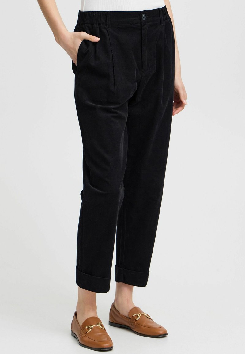 Pantaloni in corduroy neri con vestibilità rilassata, pieghe davanti, tasche laterali e orli arrotolati, abbinati a mocassini marroni.