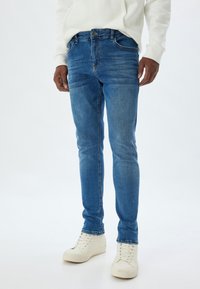 Pantalones vaqueros de mezclilla azul con un corte ajustado, que presentan un ligero desgaste, cinco bolsillos y cierre con botón en la parte delantera, combinados con zapatillas blancas.
