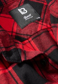 Tela de camisa de franela a cuadros rojo y negro con una textura tejida, que cuenta con una etiqueta negra con texto blanco indicando la marca y la talla.
