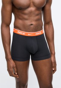 Homme athlétique portant un boxer Nike noir avec une ceinture orange arborant le logo Nike blanc et le swoosh, debout devant un fond uni.