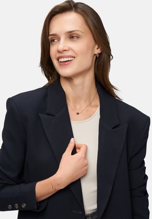 Blazer marine avec des revers structurés sur un haut beige clair. Collier et bracelet en forme de cœur doré. Cheveux lisses encadrent le visage.