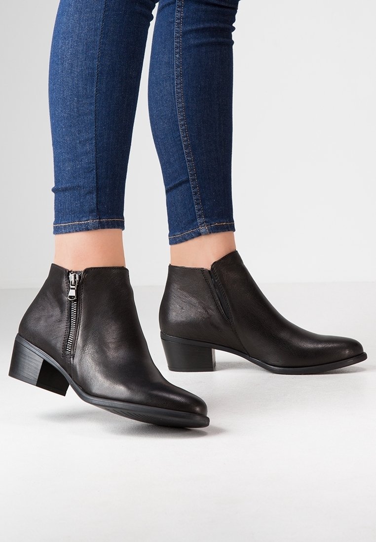 Personne portant un jean skinny bleu foncé et des bottines en cuir noir à talons blocs avec fermetures éclair latérales, debout sur un sol clair.
