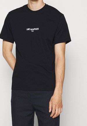 Mężczyzna ubrany w czarną koszulkę z krótkim rękawem z białym nadrukiem "THE KOOPLES PARIS" na piersi, w zestawie z ciemnymi spodniami.