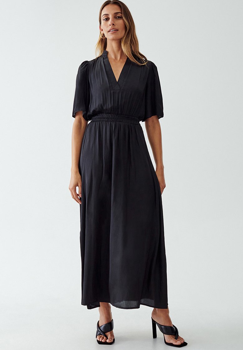 Vestido largo negro con escote en V, mangas cortas abullonadas y cintura elástica. Textura suave con una silueta fluida. Llevado con tacones negros.