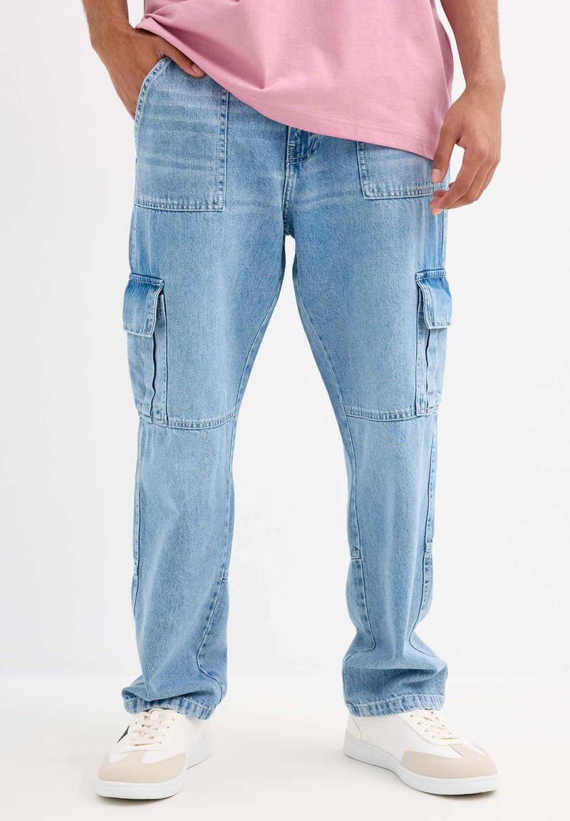 Jeans cargo de color azul claro con múltiples bolsillos grandes, un corte relajado y una textura de denim desgastado, combinados con zapatillas blancas y una camiseta rosa.