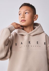 Smėlio spalvos džemperis su kapišonu, rankovėse ir apačioje su siaurėjančios tekstūros raštu. Ant priekinės dalies didelė juoda užrašas „BAKER“ ir „by TED BAKER“.