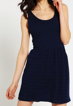 Nainen pukeutuneena hihattomaan laivastonsiniseen silmäkuviolliseen mekkoon, jossa on istuva vyötärö ja pyöreä pääntie, poseeraa yhdellä kädellä lähellä kasvojaan.