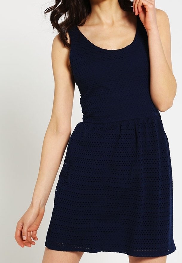 Femme portant une robe sans manches bleu marine ajourée avec une taille cintrée et un décolleté en goutte d'eau, posant avec une main près du visage.