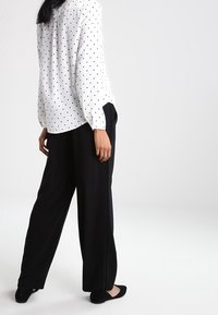 Una donna sta in piedi indossando una blusa bianca a maniche lunghe con pois neri, pantaloni larghi neri e scarpe basse nere.
