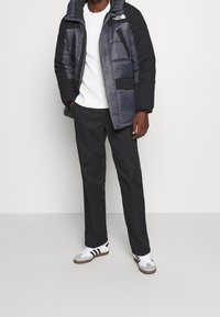 Kurtka pikowana w ciemnoszarym i czarnym kolorze z kapturem, z przednim zamkiem błyskawicznym i bocznymi kieszeniami. Nosi się ją na białym prążkowanym swetrze i ciemnych spodniach.