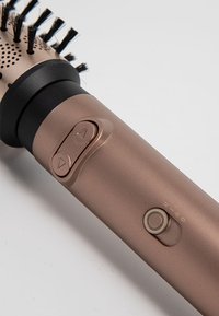 Outil de coiffure rond en bronze avec des poils, des boutons de contrôle et une poignée lisse. Accents texturés près de la tête de la brosse et de l'interrupteur marche/arrêt.