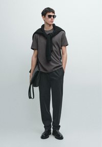 Graues T-Shirt, schwarzer Pullover am Hals gebunden, schwarze Hose und schwarze Stiefel. Accessoires: schwarze Sonnenbrille und eine Schultertasche.