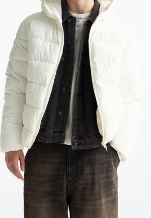 Personne portant une doudoune blanche par-dessus une veste en jean noire, avec un pantalon foncé et un collier de perles coloré visible au-dessus de l'encolure.