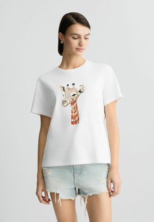 T-shirt en coton blanc avec un motif de girafe dans des tons orange et marron, à manches courtes et à coupe décontractée. Détails incluant un col rond.