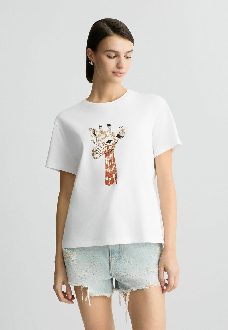 T-shirt en coton blanc avec un motif de girafe dans des tons orange et marron, à manches courtes et à coupe décontractée. Détails incluant un col rond.