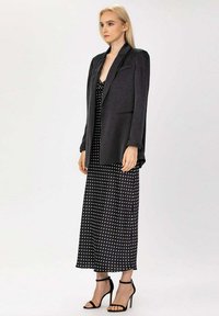 Blazer noir en tissu texturé, porté sur une robe maxi à pois noirs et blancs. La tenue est complétée par des talons noirs à lanières.