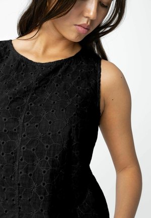 Noor naine, kes kannab musta varrukateta topi lillemustriga, vaatab alla valge tausta taustal.