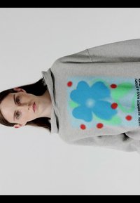 Mujer con pelo castaño liso que lleva una sudadera con capucha gris con una gran flor azul con puntos rojos y hojas verdes en el frente.